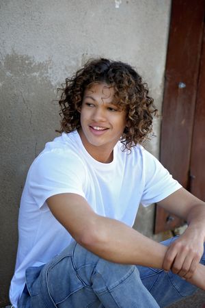 Smiling young man with curly hair, white t-shirt and blue jeans sits casually against a weathered wall and looks aside.