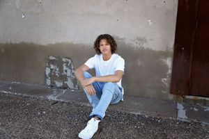 Young man in a white t-shirt, jeans and white sneakers sits on a curb in front of a weathered gray wall and rust-red door.