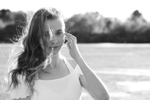 Black and white portrait of a young woman in a white top with windblown wavy hair holding her hand to her ear near water.