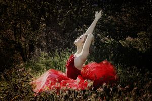 Dancer in a red tulle dress stretches one arm skyward while seated on grass in a sunlit wooded area.