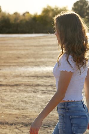 Young woman in a white smocked top and blue jeans walks along a sunlit dry lakebed with trees glowing in the background.