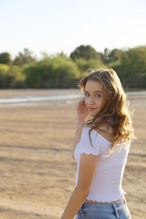 Young woman with long wavy hair looks over her shoulder in a white top and jeans at a sunlit lakeshore.