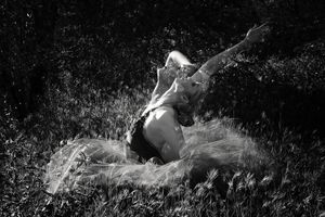 Black and white image of a dancer in a tulle skirt stretching one arm outward while reclining on sunlit grass.