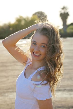 Smiling young woman with curly blonde hair raises her hand to her head in golden sunlight near a lake.