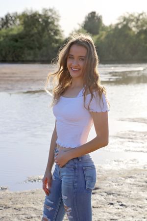 Smiling young woman in a white ruffled top and ripped jeans stands on a sandy shoreline at sunset.