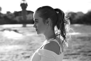 Black and white profile portrait of a young woman with a ponytail standing by the water in soft backlight.