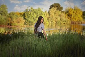 Young woman with long dark hair walks through tall green grass toward a glowing lake with golden autumn trees in the background.