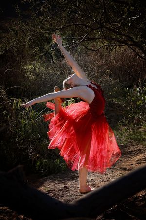 Barefoot ballet dancer in a flowing red tutu dress arches backward with arms extended on a woodland trail.