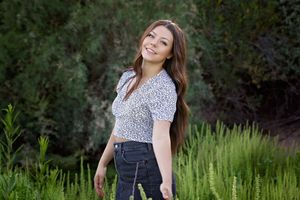 Smiling young woman in a patterned crop top and dark jeans poses in a green grassy field with trees in the background.