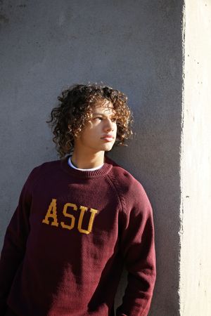 Young man with curly hair wearing a maroon ASU sweater leans against a concrete wall while gazing off into the distance.