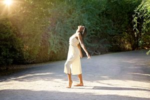 Barefoot woman in a flowing white dress dances along a sunlit path lined with green trees as the sun glows through the leaves.
