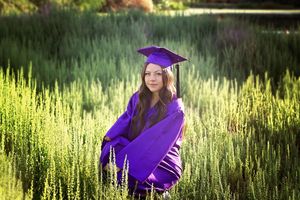 Smiling graduate in a purple cap and gown poses among tall green grass in warm golden light for a senior portrait.