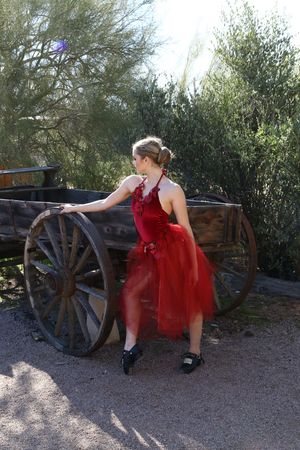 Dancer in a red tulle dress rests her hand on an antique wooden wagon wheel with desert trees and bright sunlight behind her.