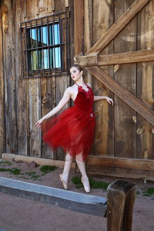 Ballerina in a red tutu dress and pointe shoes poses gracefully beside a weathered wooden barn with a barred window.