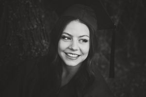 Black and white portrait of a smiling graduate in a cap looking to the side with long dark hair against a moody background.