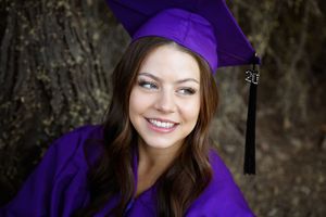 Smiling young woman in a purple graduation cap and gown with a tassel poses in front of a tree trunk for a graduation portrait.