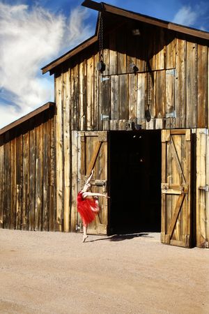 Ballerina in a red tutu performs a leg extension in front of a rustic wooden barn with open doors under a blue sky with clouds.