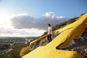 Person in a white shirt stands on a yellow-painted rocky hillside overlooking a city under a bright blue sky with large clouds.