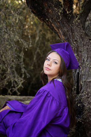 Young woman in a purple graduation cap and gown leans against a tree trunk in a wooded outdoor setting for her senior portrait.