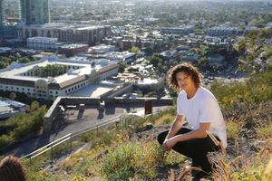 A young man with curly hair crouches on a grassy hilltop overlooking a sunlit city with buildings below.