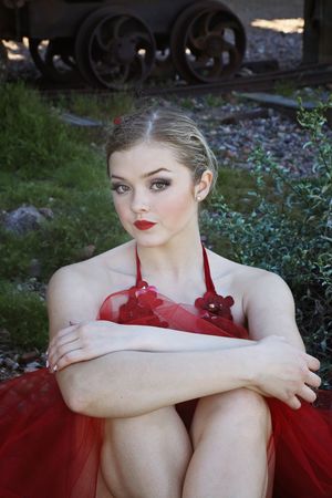 A blonde ballerina with red lipstick and a red tulle costume sits with arms around her knees near old rail wheels.