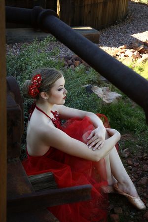 A ballerina with a red flower in her hair sits beside weathered wooden beams, wearing a red tulle dress and pointe shoes.