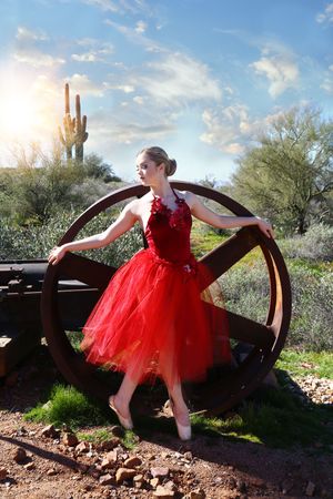 A ballerina in a red tutu poses en pointe inside a large wooden wheel with a saguaro cactus and sunlit sky behind her.