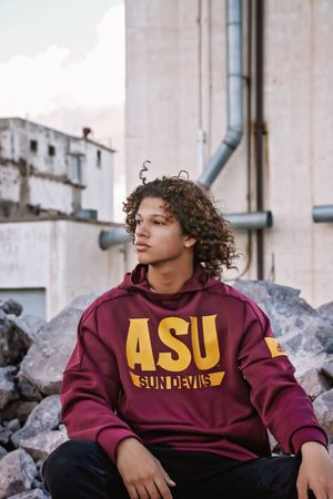 A young man with curly hair wears a maroon ASU Sun Devils hoodie while sitting on rocks in front of an industrial building.