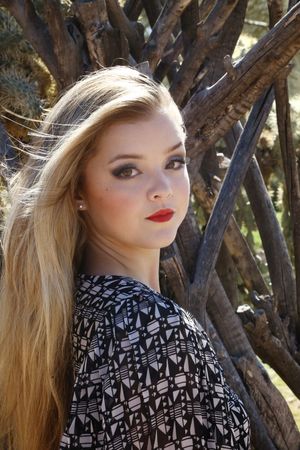 Young woman with long blonde hair and red lipstick wearing a geometric print top poses beside weathered wooden branches.