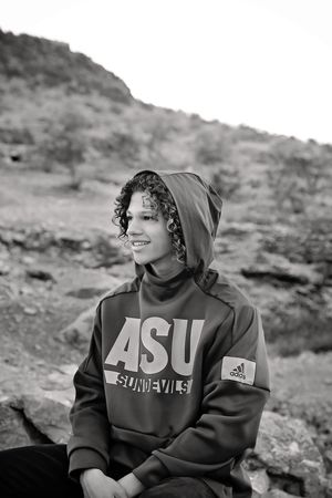 Black and white photo of a smiling young person with curly hair in an ASU Sun Devils hooded sweatshirt sitting on desert rocks.