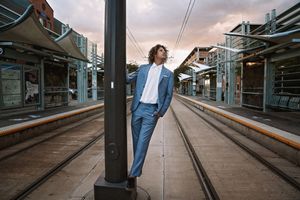 A curly-haired man in a blue suit leans against a pole on a train platform with tracks stretching into the evening light.