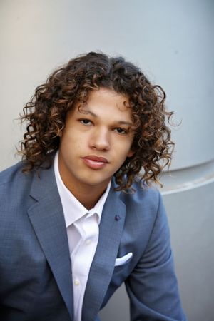 Portrait of a young man with curly brown hair wearing a blue blazer and white shirt with a pocket square against a gray wall.