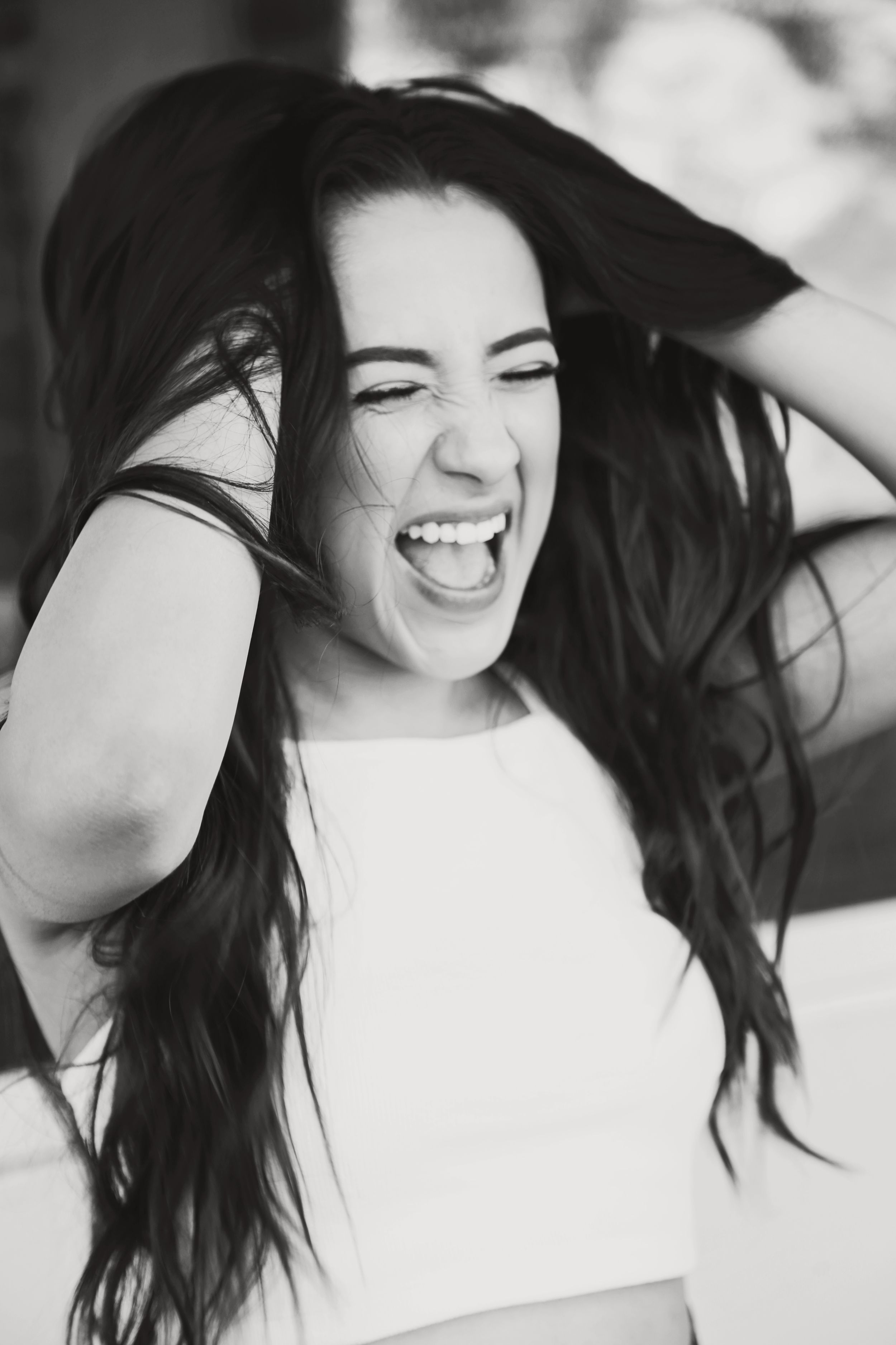 Black and white portrait of a woman with long dark hair laughing joyfully with her hands in her hair.