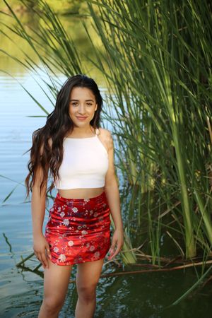 Smiling young woman in a white crop top and red floral skirt standing in shallow water beside tall reeds.