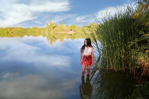 Woman in a white top and red skirt wading in a calm lake with reeds, blue sky and reflected clouds behind her.