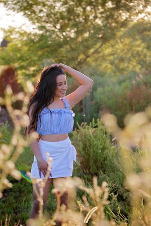 Smiling woman in a blue ruffled top and white skirt posing with a hand on her head in a sunlit garden.