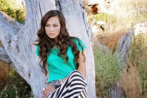 A young woman with long wavy hair and a teal top sits against a weathered driftwood tree trunk in a dry grassy field.
