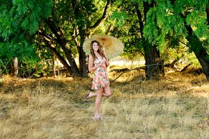 A smiling girl in a floral dress holds a bamboo parasol while posing on one leg in a sunlit grassy woodland.