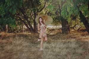 Young woman in a floral dress twirls a yellow parasol on a dry grassy field among tall trees in soft sunlight.
