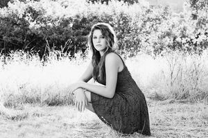 Black and white portrait of a young woman in a lace dress crouching in a tall grass field with soft backlight.