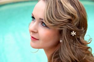Close-up profile of a woman with wavy hair, a floral hair clip, and pearl earring beside a bright turquoise swimming pool.