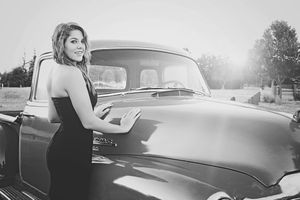 Black and white photo of a woman in a fitted dress leaning on a vintage pickup truck hood at sunset.