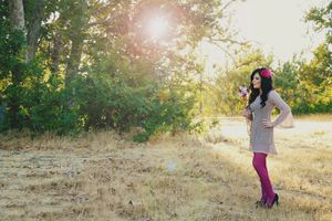 Woman in a gray dress and magenta tights stands in a sunlit golden field holding flowers, with trees glowing in the background.
