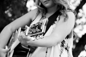 Black and white close-up of a woman in a fringed top playing an acoustic guitar outdoors with soft sunlight behind her.