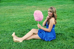 Smiling woman in a blue dress sits on lush green grass holding a large pink cotton candy on a stick.