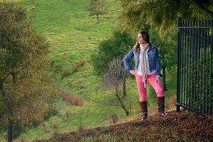 Young woman in a denim jacket and pink pants poses on a grassy hillside beside a black iron gate at golden hour.