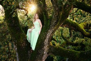 Woman in a long mint green dress sits on a mossy oak tree trunk in an enchanted forest lit by warm sunlight.