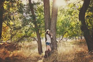 Woman in a cowboy hat and denim shorts leans against a tree trunk in a sunlit woodland with warm autumn tones.