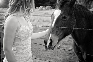 Black and white photo of a woman in a lace dress gently petting a horse through a barbed wire fence.