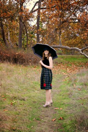 Smiling woman in a plaid skirt holds a black umbrella on a leaf-strewn path surrounded by orange autumn trees.
